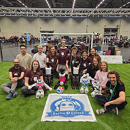 RoboCup Team der HU "Berlin United" Ein Team posiert mit Robotern auf einem Kunstrasenfeld bei der RoboCupHumanoid Soccer League.
