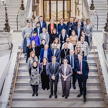 Große Gruppe von etwa dreißig Personen steht für ein Gruppenfoto auf einer monumentalen steinernen Treppe in einem historischen Gebäude mit hohen Decken, Bögen und reich verzierten Wänden. Mehrere antike Lampen flankieren die Treppe, und im Hintergrund öffnen sich weitere Treppenaufgänge und ein großer Torbogen.