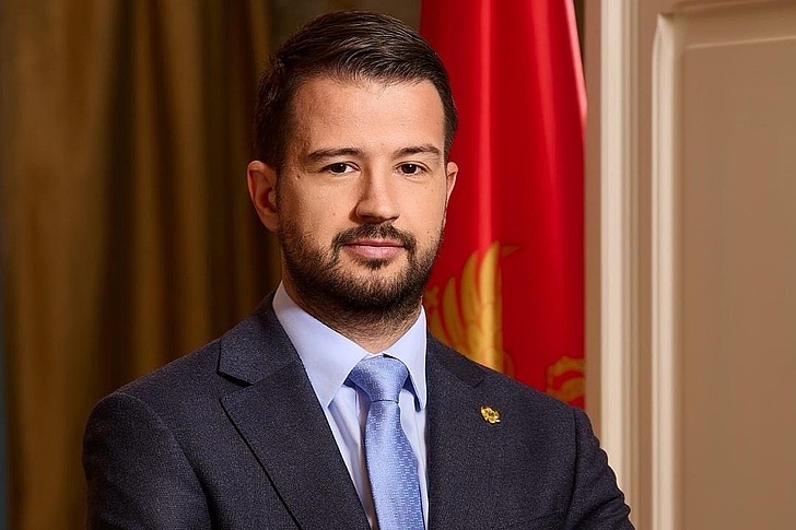 Jakov Milatović Jakov Milatović in Anzug und Krawatte mit Bart, vor einer Flagge und Vorhängen.