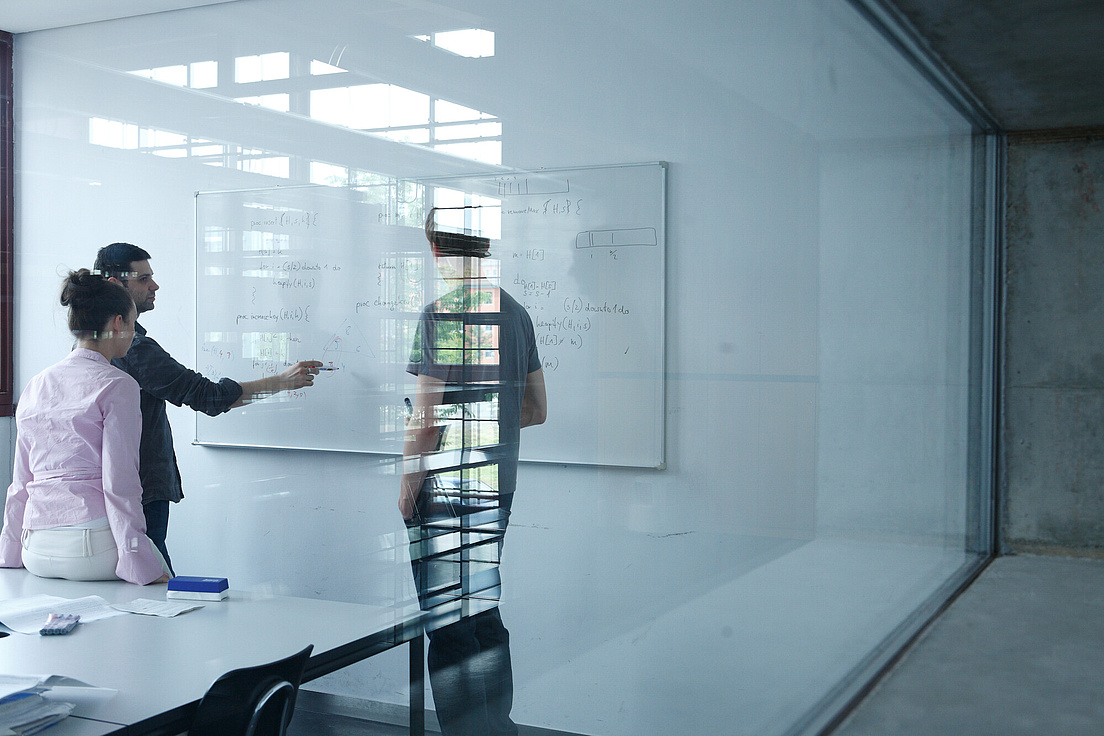 Studierende des Campus Adlershof Das Bild zeigt den Blick durch eine große Scheibe, dahinter befindet sich ein Whiteboard, auf der verschiedene mathematische Formeln stehen. Vor der Tafel stehen drei Menschen, wovon eine Person auf dem Tisch vor der Tafel sitzt und eine andere scheinbar erklärend mit dem Stift auf die Tafel zeigt.