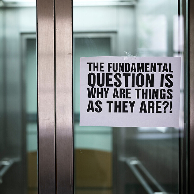 Eine Glastür eines geschlossenes Aufzuges. Darauf klebt mit Tesafilm ein Schild auf dem steht The fundamental Question ist, why are things as they are?