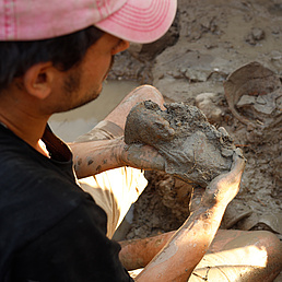 A man wearing a pink cap holds a mud-covered find, while another person holds a plastic bag.