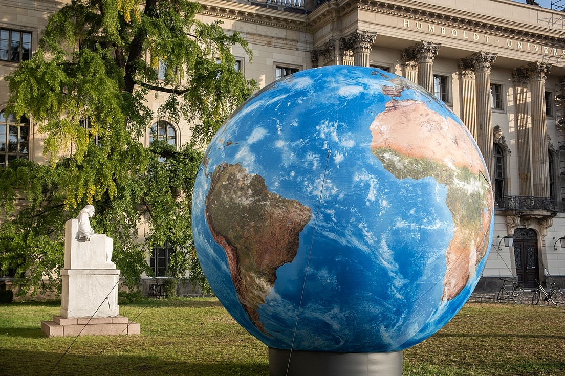 Large globe displayed on a lawn in front of the main building of Humboldt University in Berlin, next to a white statue and surrounded by trees and historic architecture.