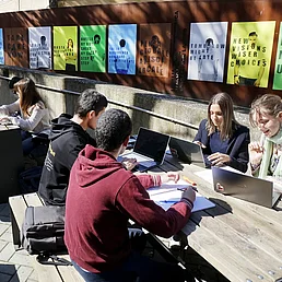 Students sit at a long wooden table outdoors working with laptops and notebooks with colorful posters on a wall in the background.