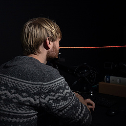 Experimental setup for an experiment conducted by the Science of Intelligence (SCIoI) Cluster of Excellence in Berlin A man wearing a patterned jumper is sitting in a dark room, watching a red laser beam.