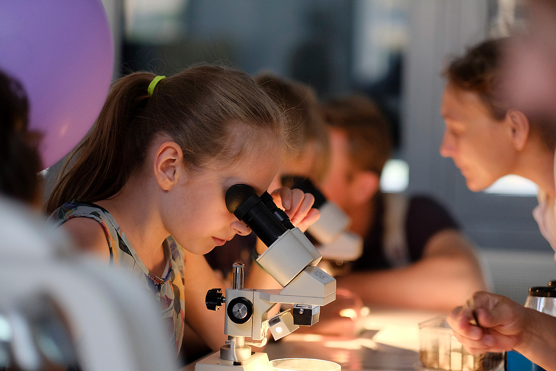 Lange Nacht der Wissenschaften Ein Mädchen schaut durch ein Mikroskop. Neben ihr sind weitere Personen, die das ebenfalls tun. Der Mikroskoptisch ist hell beleuchtet.