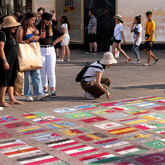 Berlin: Zahlreiche Flaggen aus Ländern weltweit liegen auf dem Boden. Eine Menschenmenge steht drum herum, eine Person kniet und schaut sie sich an, eine andere fotografiert sie.
