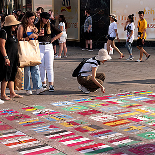 Berlin: Zahlreiche Flaggen aus Ländern weltweit liegen auf dem Boden. Eine Menschenmenge steht drum herum, eine Person kniet und schaut sie sich an, eine andere fotografiert sie.