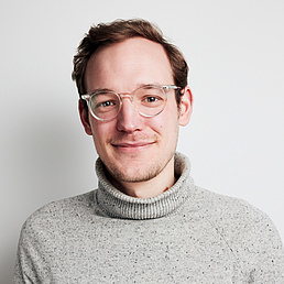 Dr. Dimitri Spieker Ein Mann mit Brille und grauem Rollkragenpullover lächelt vor weißem Hintergrund.