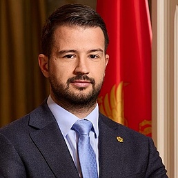 Jakov Milatović Jakov Milatović in Anzug und Krawatte mit Bart, vor einer Flagge und Vorhängen.