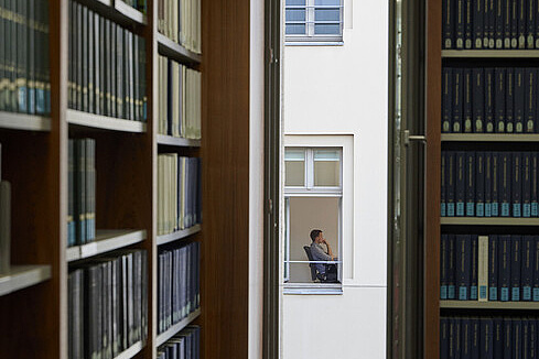 Wissenschaftliches Arbeiten Eine Person sitzt auf einem Stuhl an einem Tisch. Das Foto ist so aufgenommen, dass die Person durch ein schmales Fenster fotografiert wurde. Man sieht sie nur von der Seite im gegenüberliegenden Gebäude. Im Vordergrund sind Bücherregale aus dunklem Holz zu sehen.