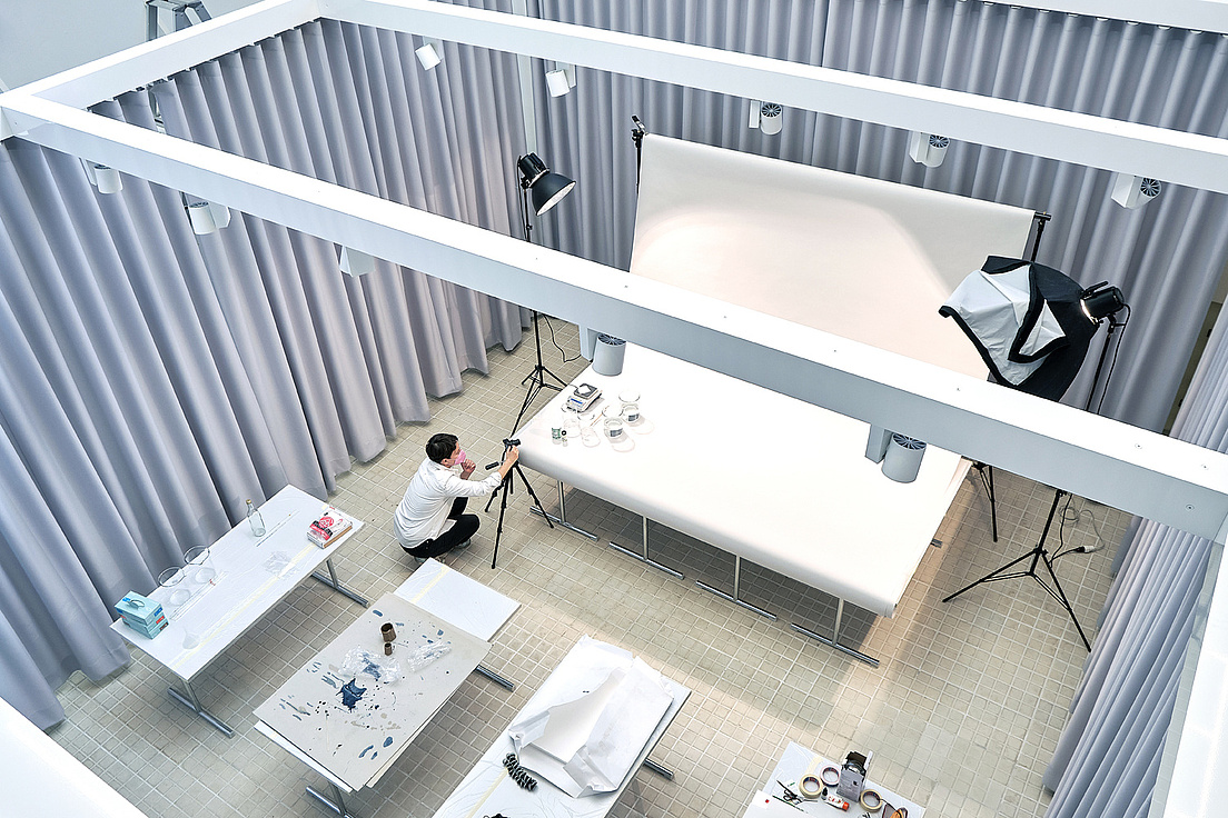 Das Bild zeigt das Objektlabor aus der Vogelperspektive. Ein Mann kniet auf dem Boden und richtet eine Kamera auf einem Stativ ein. Der Raum ist hell beleuchtet, mit weißen Tischen und verschiedenen Objekten darauf. Drum herum sind in einem Viereck graue Vorhänge gezogen.