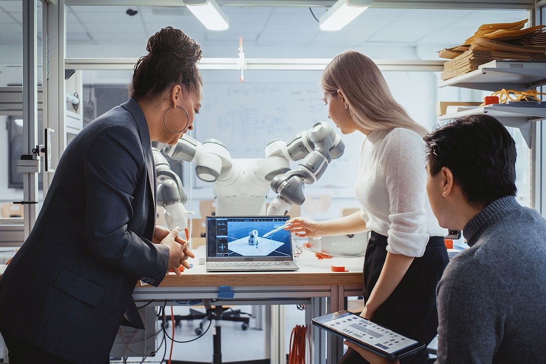 Diverse Team: Chief Engineer and Promising Computer Scientists Analyse and Discuss Optimisation of Autonomous Manufacturing Facility That Creates Great Products. Laboratory with Robotic Arm. Three people are working together in a modern laboratory or research room. They are standing around a table with a laptop, whose screen displays a 3D model of a robot. Behind them is a large industrial robot arm.
