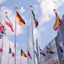 Flags of European countries wave in front of modern glass buildings under a blue sky.