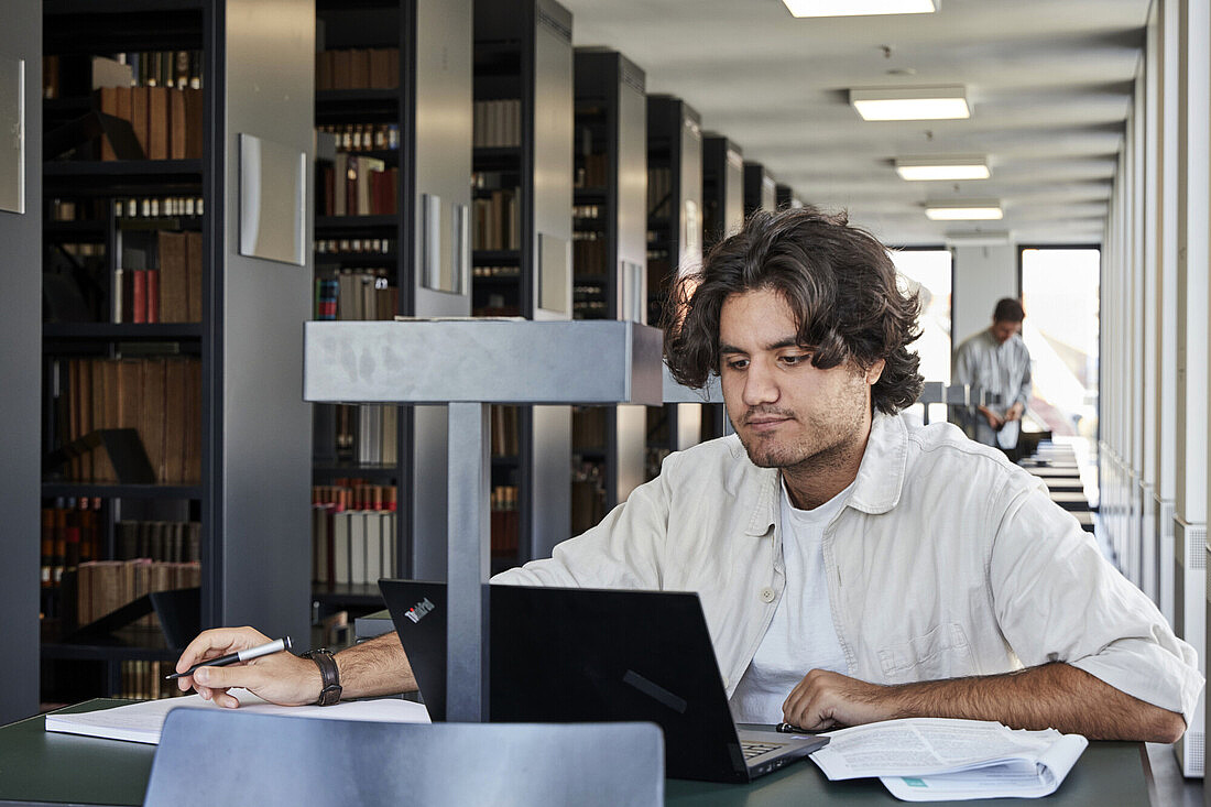 Universitätsbibliothek der Humboldt-Universität Ein junger Mann sitzt an einem Tisch in einer Bibliothek und arbeitet konzentriert an seinem Laptop. Er hält einen Stift in der Hand und hat Papiere vor sich liegen. Im Hintergrund sind Bücherregale und ein weiterer Mensch zu sehen, der ebenfalls beschäftigt ist. Die Bibliothek wirkt ruhig und gut beleuchtet.