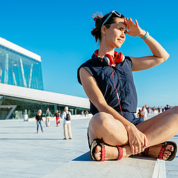 Young hipster woman traveler with red headphones resting on border after walking in Oslo city over Opera House urban background, happy, smiling, looking in sunset. Young hipster woman traveler with red headphones resting on border after walking in Oslo city over Opera House urban background, happy, smiling, looking in sunset.