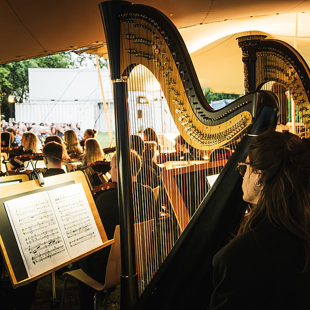 Sommeroper 2025 Konzertszene im Freien unter einem Zelt. Im Vordergrund sind zwei Harfen zu sehen, die von Musikern gespielt werden. Die Musiker sitzen auf Stühlen und haben Notenständer vor sich, auf denen Notenblätter befestigt sind. Im Hintergrund ist ein Orchester mit verschiedenen Instrumenten wie Streichinstrumenten zu erkennen. Das Publikum sitzt auf Stühlen und hört dem Konzert zu.