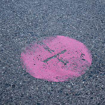 Das Bild zeigt einen rosa Kreis, der auf einer asphaltierten Oberfläche mit einem schwarzen Pinselstrich markiert ist. Der Hintergrund besteht aus grauem Asphalt mit kleinen Steinen und Kieseln.