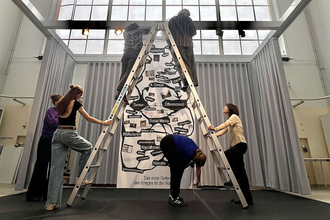 Open Humboldt, Zentrum für Kulturtechnik Drei Personen stehen auf einer Leiter, vor einer Plakatwand. Eine vierte Person bringt etwas auf der Wand an.