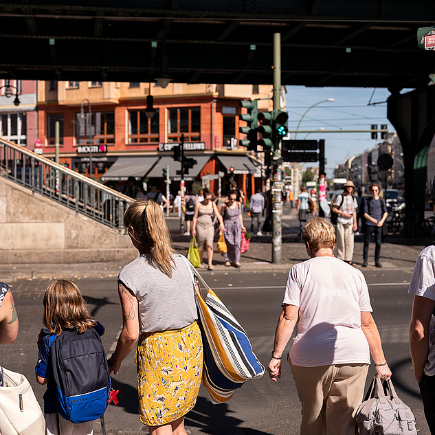 Open-Humboldt-Gesundheit-Berlin-Stadtansichten-Matthias-Heyde Das Bild zeigt eine belebte Straßenszene in einer Stadt. Mehrere Menschen sind zu sehen, die entweder auf der Straße stehen oder darauf warten, sie zu überqueren. Im Vordergrund befinden sich fünf Personen, darunter eine Frau mit einem Kind, das einen Rucksack trägt. Im Hintergrund sind weitere Fußgänger zu sehen, die sich auf der Straße bewegen. Es gibt auch einige Geschäfte und Cafés mit Schildern.