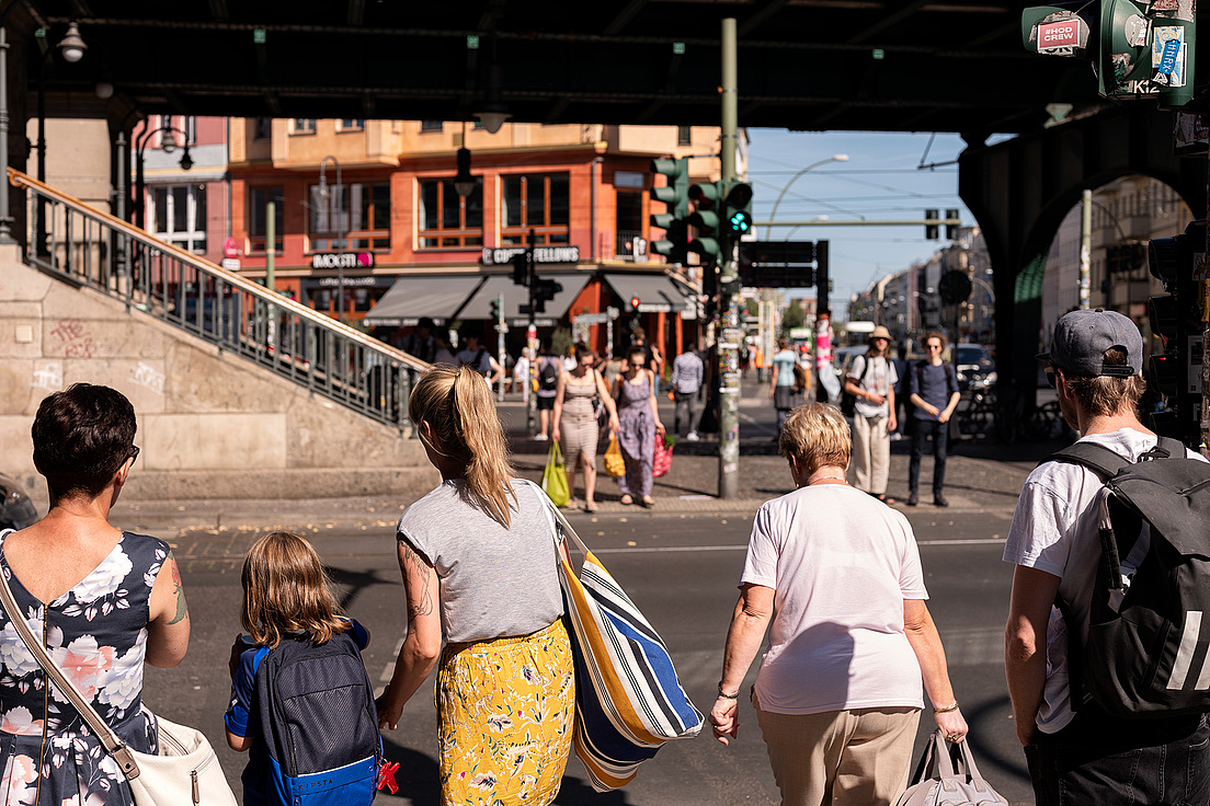 Open-Humboldt-Gesundheit-Berlin-Stadtansichten-Matthias-Heyde Das Bild zeigt eine belebte Straßenszene in einer Stadt. Mehrere Menschen sind zu sehen, die entweder auf der Straße stehen oder darauf warten, sie zu überqueren. Im Vordergrund befinden sich fünf Personen, darunter eine Frau mit einem Kind, das einen Rucksack trägt. Im Hintergrund sind weitere Fußgänger zu sehen, die sich auf der Straße bewegen. Es gibt auch einige Geschäfte und Cafés mit Schildern.