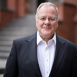 Georg Essen Georg Essen trägt ein weißes Hemd und einen dunkelfarbenen Anzug. Er steht vor dem Eingangsportal des Zentralinstituts für Katholische Theologie der HU. Es ist ein rotfarbenes Backsteingebäude.