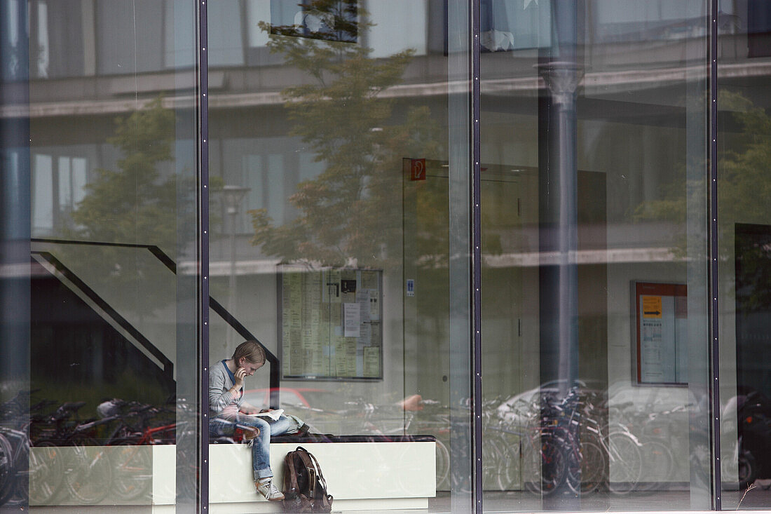 Das Bild zeigt eine Person, die in einem Innenraum an einer Wand sitzt und ein Buch liest. Die Szene wird durch eine Glasfassade betrachtet, die Reflexionen von Bäumen, Fahrrädern und einem Gebäude zeigt. Neben der Person steht ein Rucksack auf dem Boden. Im Hintergrund sind Tafeln mit Informationen zu sehen.