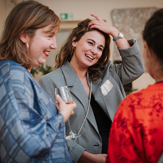 Vernetzungsevents der Humboldt-Innovation GmbH Drei Frauen unterhalten sich bei einer Veranstaltung. Die Frau in der Mitte, in einer karierten Jacke, lacht herzlich und berührt ihr Haar. Die Frau links von ihr, in einer blauen Jacke, hält ein Glas und lacht ebenfalls. Die dritte Frau, rechts im Bild, trägt ein rotes Oberteil mit einem Muster und ist dem Gespräch zugewandt. Im Hintergrund ist eine Projektion zu sehen.