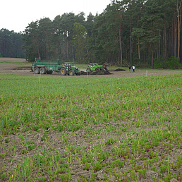 Dünger aus menschlichen Ausscheidungen Zu sehen ist ein Feld im Hintergrund ist eine landwirtschaftliche Maschine zu sehen, die den Dünger aus menschlichen Ausscheidungen auf dem Feld verteilt. Im Hintergrund ist der Anfang eines Waldes zu sehen.