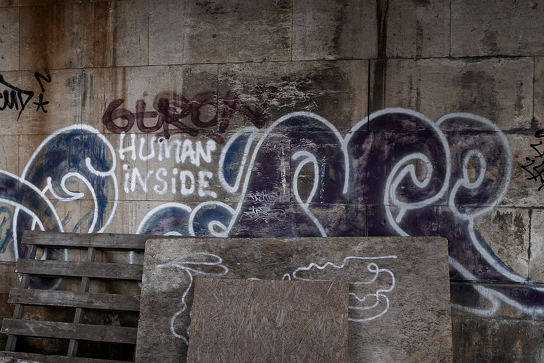 Das Bild zeigt eine Betonwand mit Graffiti, auf der die Worte "HUMAN INSIDE" in weißer Farbe zu lesen sind. Darüber und daneben sind abstrakte blaue und violette Muster. Vor der Wand stehen ein Holzpalette, eine Holzplatte und ein Sperrholzbrett.