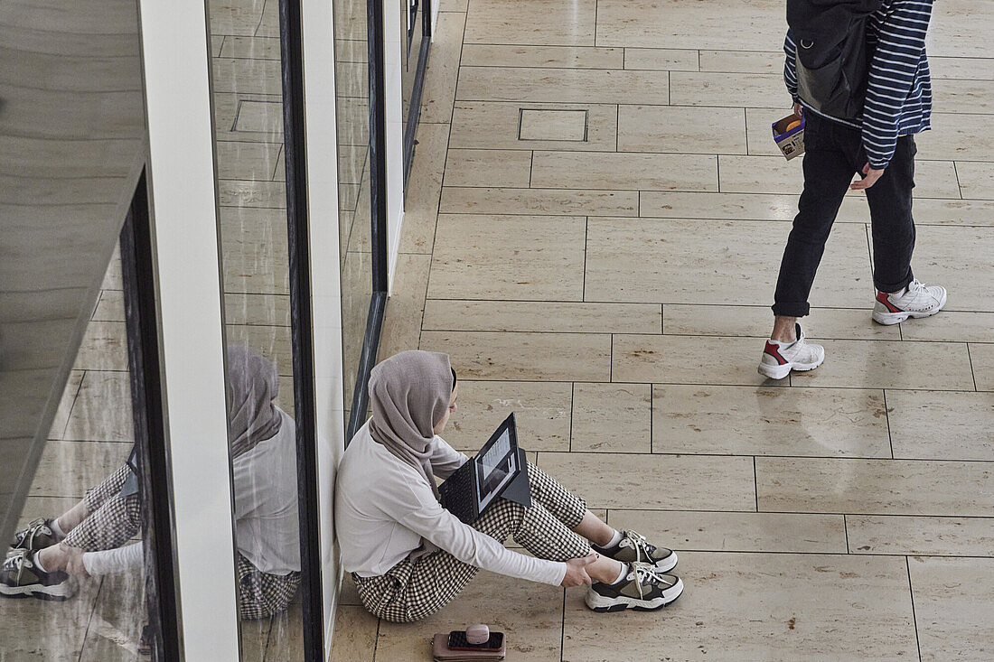 Eine Frau sitzt mit ihrem Laptop auf dem Boden eines Gebäudes mit hohen gläsernen Türen. Sie hat ihren Laptop auf den Knien liegen und schaut sich darauf etwas an.