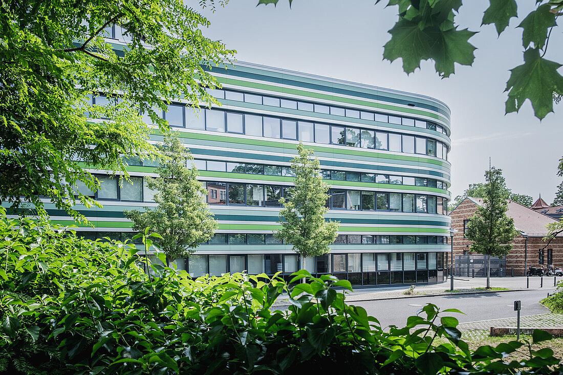 Campus Nord Rhoda-Erdmann-Haus mit vielen Fenstern, runder Gebäudeform und einer breiten Straße davor. Zu sehen sind auch viele Bäume und Gebüsche rund um das Gebäude.