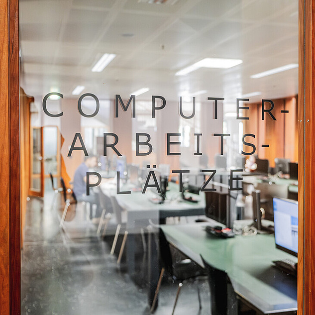 Arbeitsplätze im Grimm-Zentrum Eine Glastür, die in hellbraunes Holz eingelassen ist. Im Hintergrund sieht man Arbeitsplätze. Auf der Tür steht Computer-Arbeitsplätze